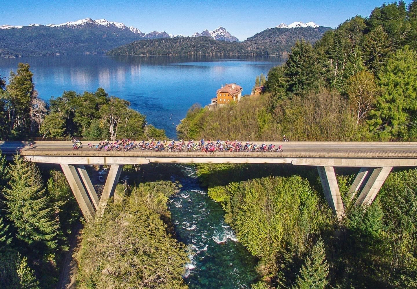 Pelotón de ciclistas cruzando un puente sobre un río, con lago y cordillera